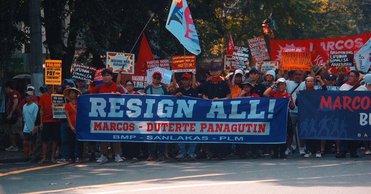 Crowd gathers in street protest holding signs demanding resignation with political messages.