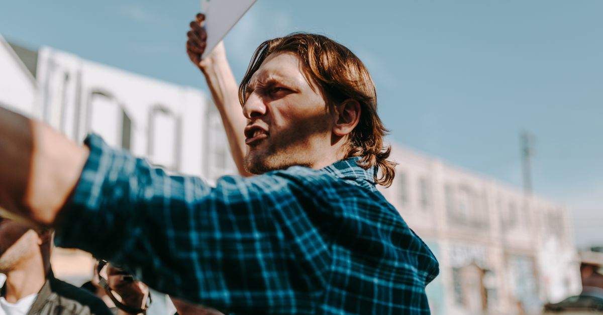 Man in a plaid shirt passionately speaking and holding a sign during an outdoor protest.