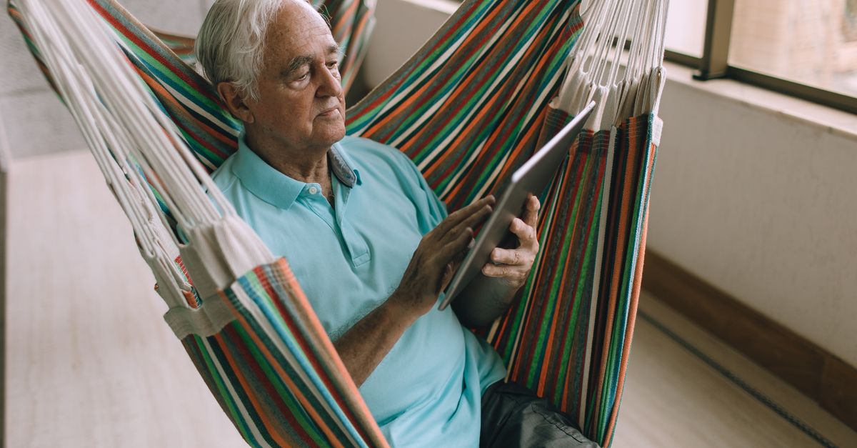 Elderly man enjoying leisure time indoors in a hammock, using a tablet for reading or browsing.
