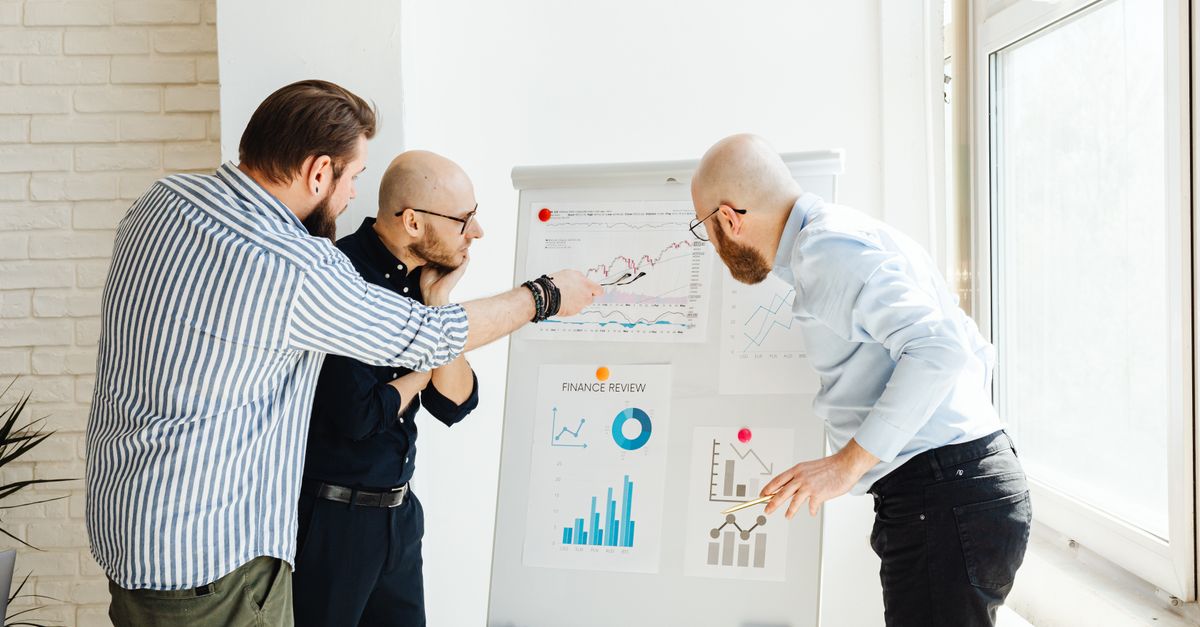 Three colleagues in a bright office discuss financial charts on a whiteboard.