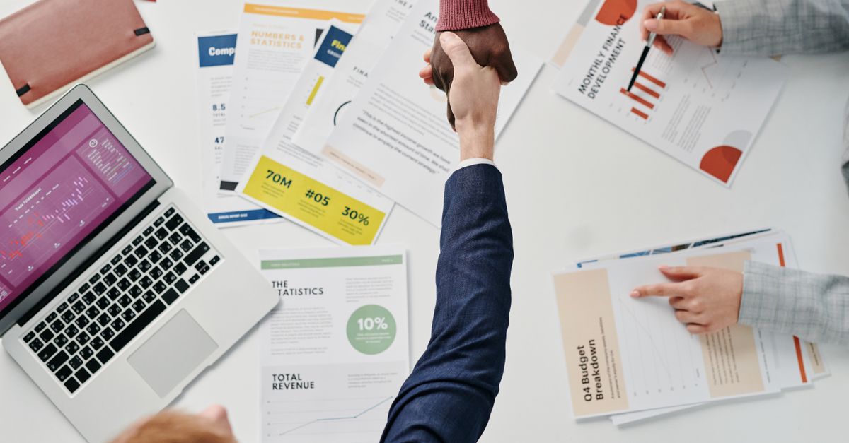 Business professionals engaging in a handshake over a desk filled with financial documents and a laptop.