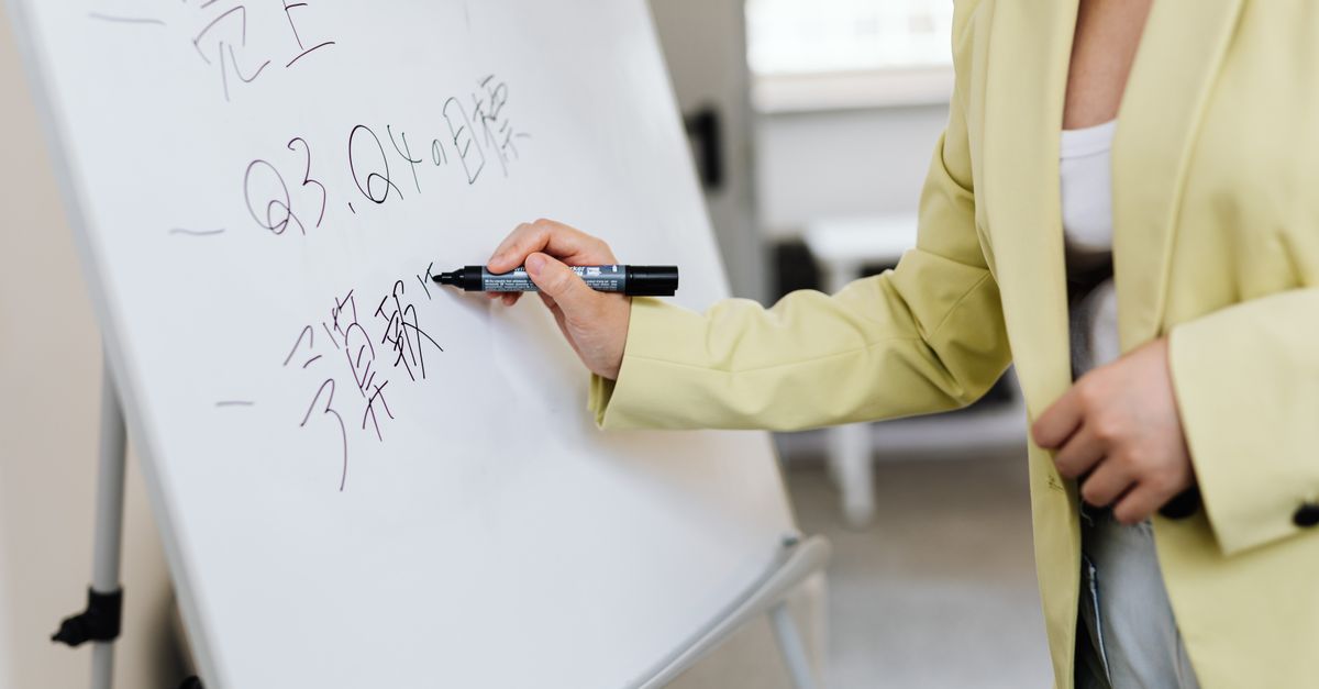 East Asian woman writing on a whiteboard during a business presentation with Chinese characters visible.
