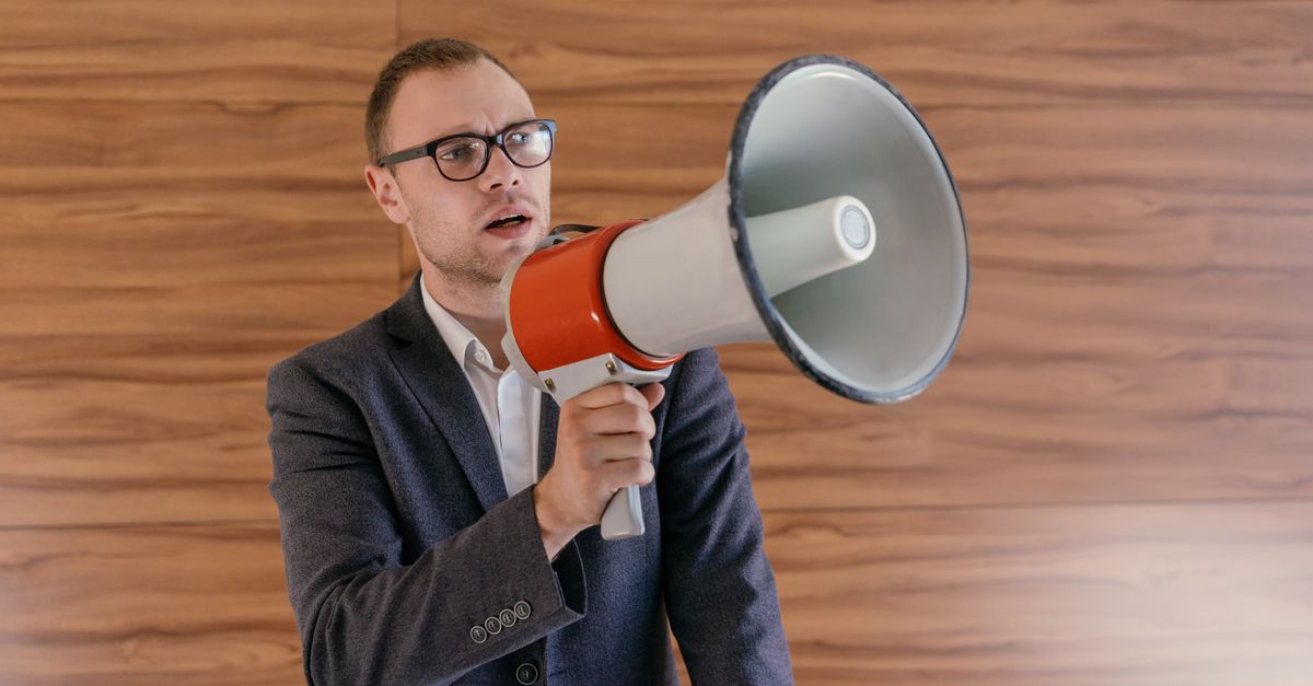 Man in suit holding a megaphone, making a public announcement indoors.