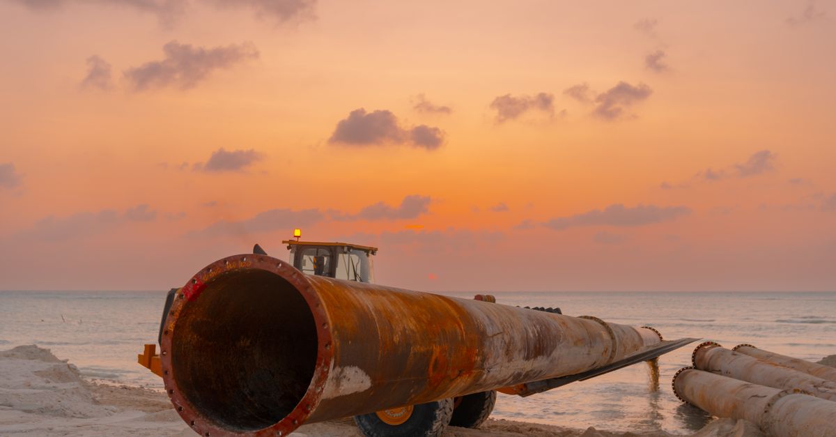 A scenic beach construction site at sunset with heavy machinery and large pipes.
