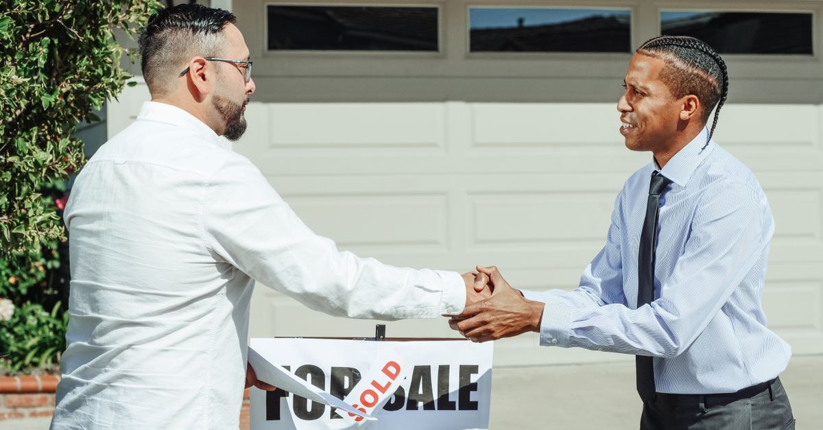 Two men shaking hands in front of a sold house sign, sealing a real estate transaction.