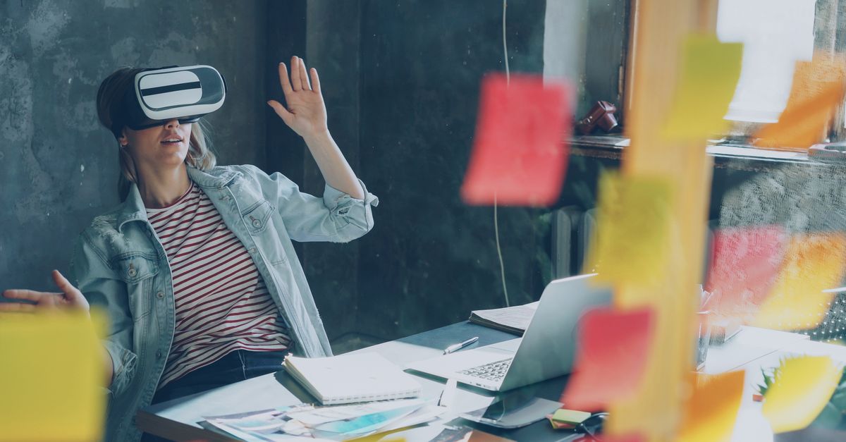 Woman using VR headset at desk, brainstorming with sticky notes and laptop.