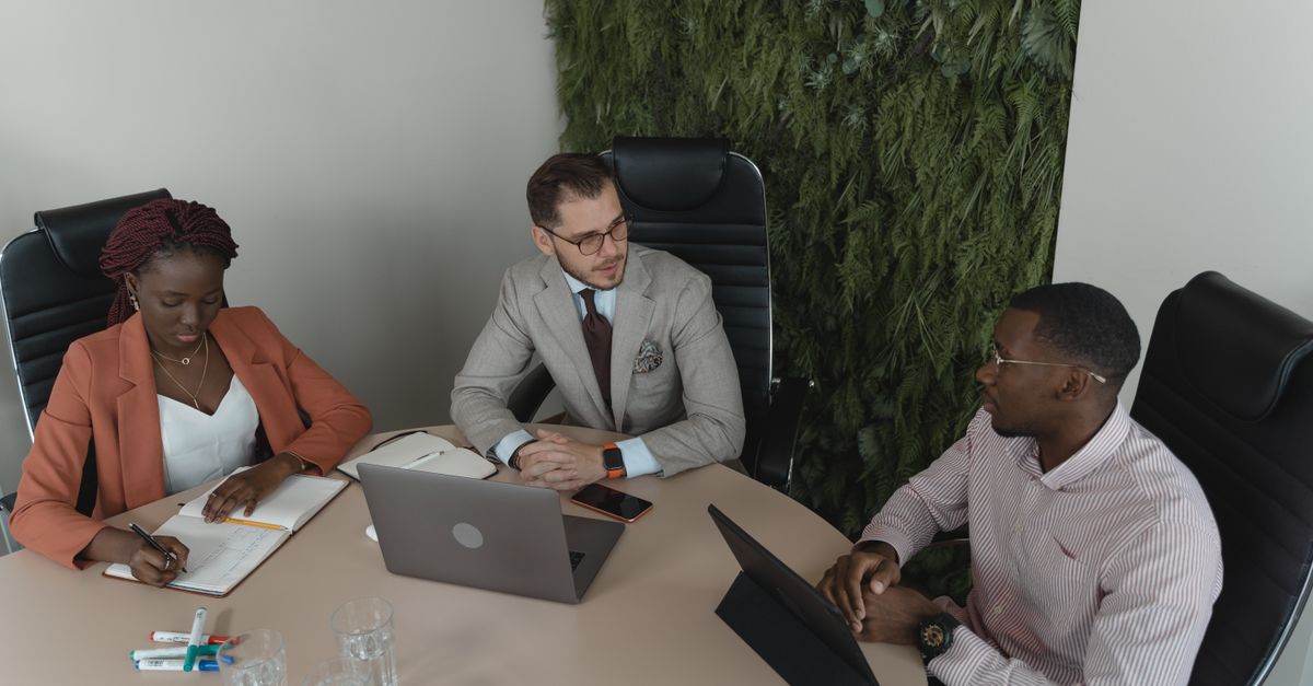 Three professionals engaged in a collaborative office meeting with laptops and notes.